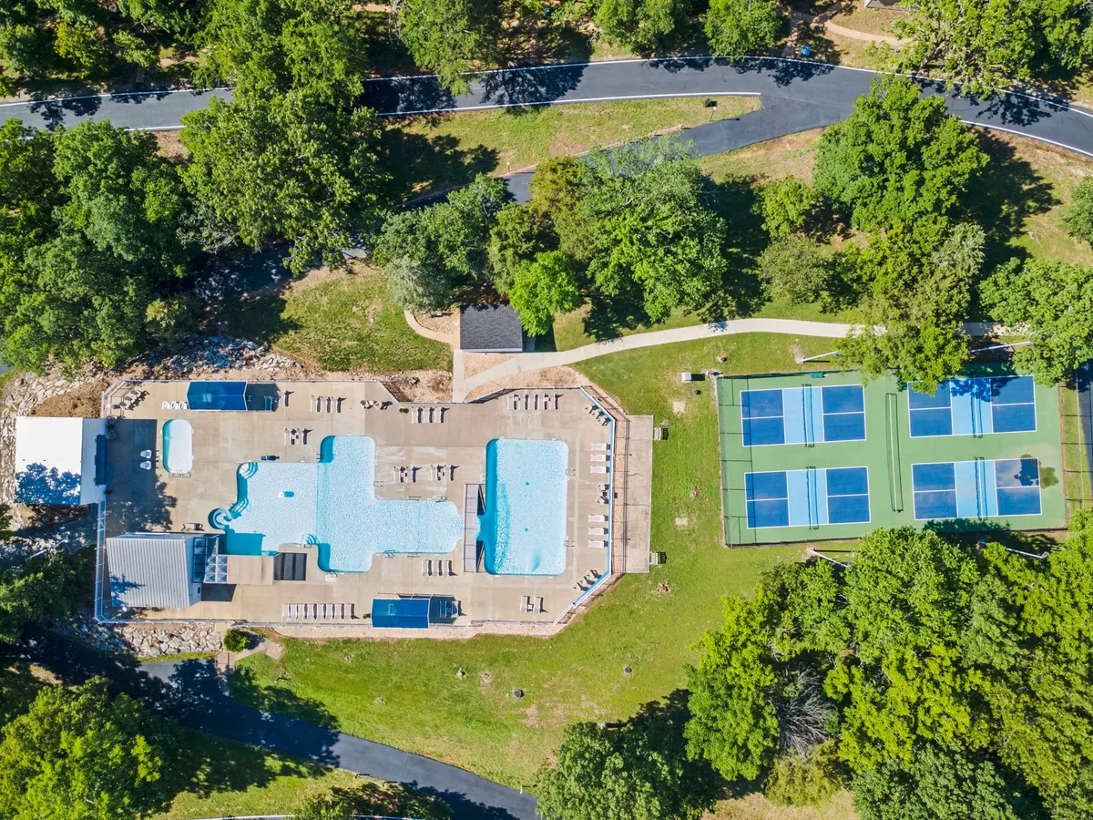 A sparkling blue swimming pool flanked by tennis courts and surrounded by green trees, with people enjoying swimming, tennis, and relaxing by the pool. The scene exudes a leisurely, community-oriented mood.