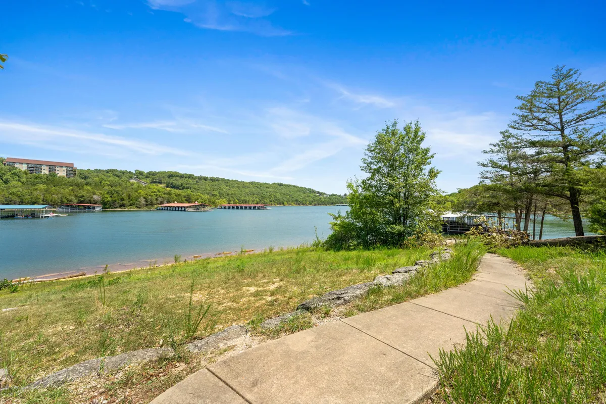This image captures a tranquil lakeside scene, with a clear pathway winding beside the water, surrounded by trees, hills, and nearby boathouses. The setting radiates peace and natural beauty, and the pathway suggests easy access for walks along the lake.