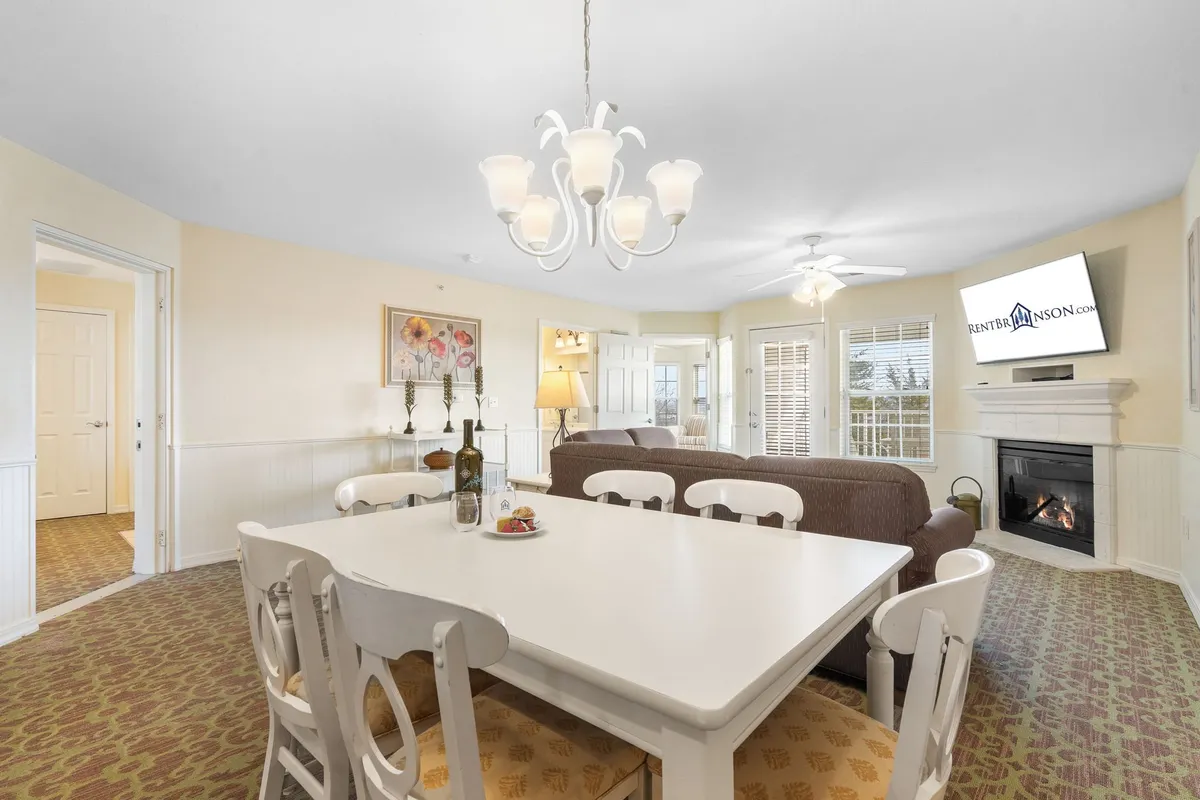 This image captures a warm, open-concept living and dining area. A dining table sits beneath a glowing chandelier near a plush sofa and a cozy fireplace, with a television positioned for easy movie nights. Large windows allow natural light to pour in, helping the space feel bright and inviting.