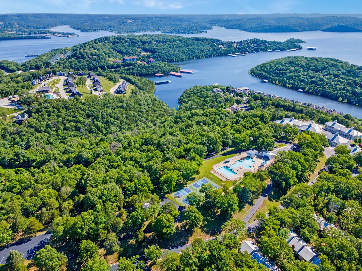 An inviting panoramic view of a resort community shows a sparkling lake, a swimming pool, tennis courts, lush greenery, and residential buildings. The open outdoor spaces feel welcoming and active, with the lake as the central focal point.