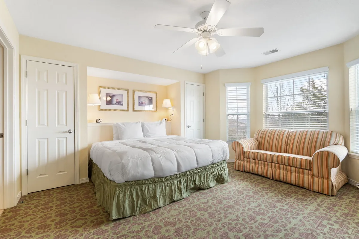 This image features a king-sized bed with plush bedding, a striped couch, framed artwork, and windows that allow natural light to fill the room. The soothing color palette and comfortable furnishings create a tranquil, inviting atmosphere ideal for relaxation.