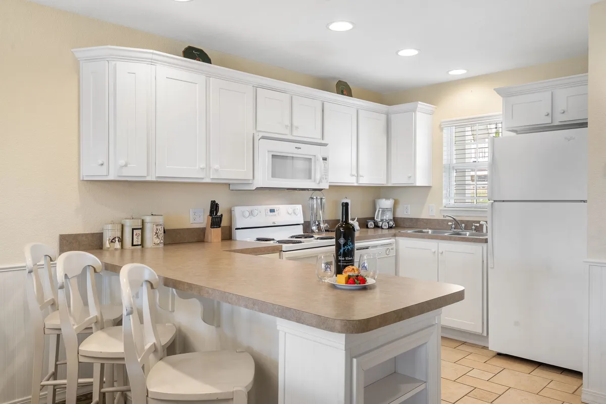 The image showcases a bright, inviting kitchen with a central island adorned with wine and fresh fruit. Crisp white cabinetry and modern appliances create a clean, homey atmosphere perfect for gathering, cooking, and making memories.