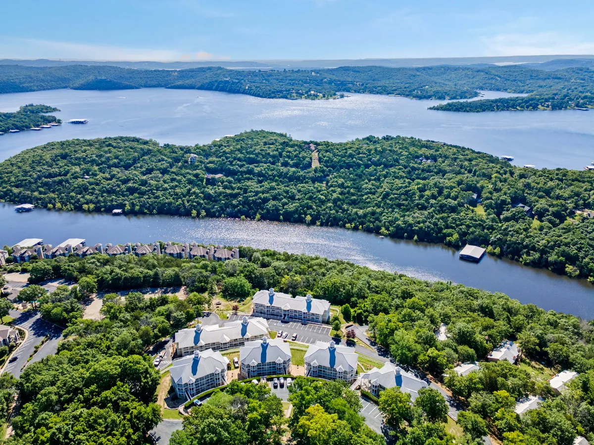 A wide-angle view of Table Rock Lake, surrounded by rolling green hills and residential buildings. The scene is tranquil, with calming water and a hint of sunset. A boathouse and winding roadways invite exploration and relaxation by the lake.