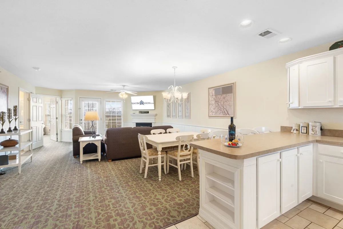 A welcoming interior scene showing a dining table adjacent to a sofa, with a chandelier above and the kitchen counter nearby. The space is filled with natural light, creating an inviting setting for shared meals and relaxed conversation.