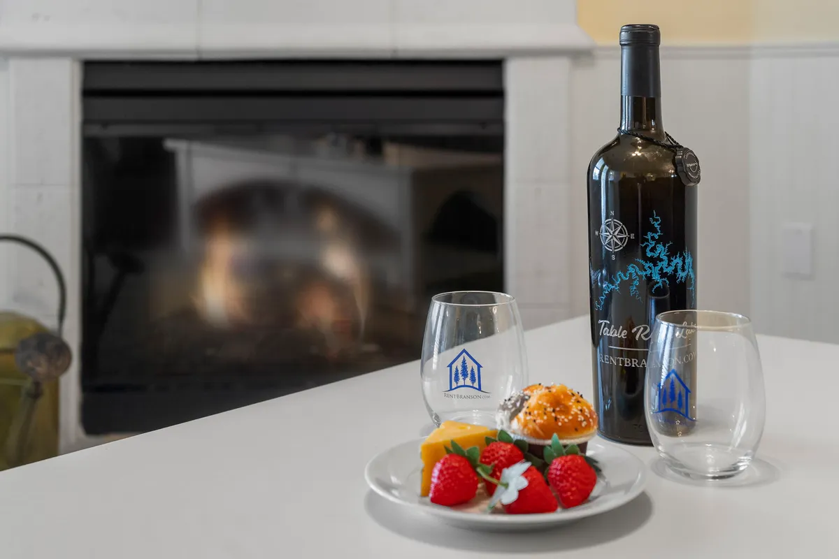 A cozy interior scene featuring a wine bottle, dessert plate, glasses, and a crackling fireplace. The inviting arrangement suggests a warm, intimate gathering with friends or a romantic evening indoors.