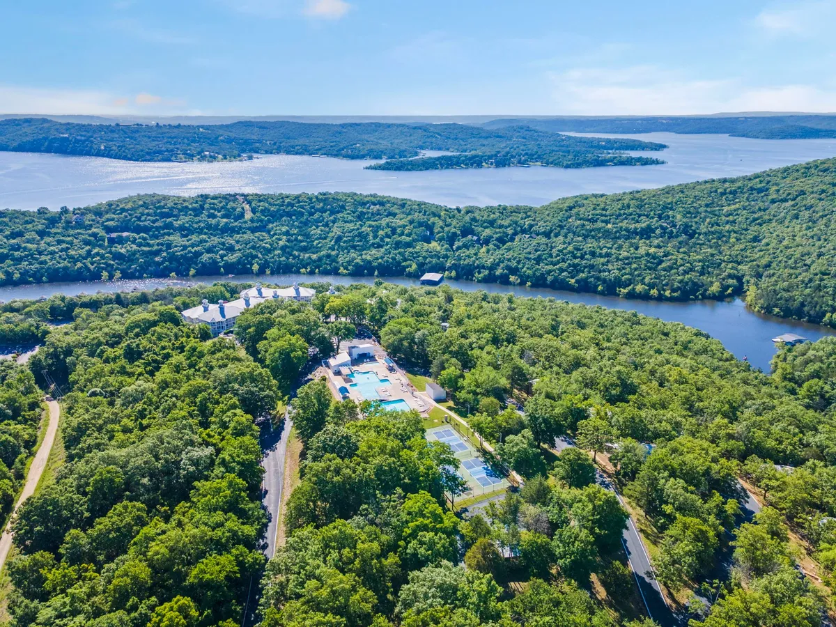 An elevated view showing lush green trees surrounding sparkling blue water, with a swimming pool and tennis courts in the foreground. The image highlights both natural and recreational amenities, with clear skies and inviting facilities.