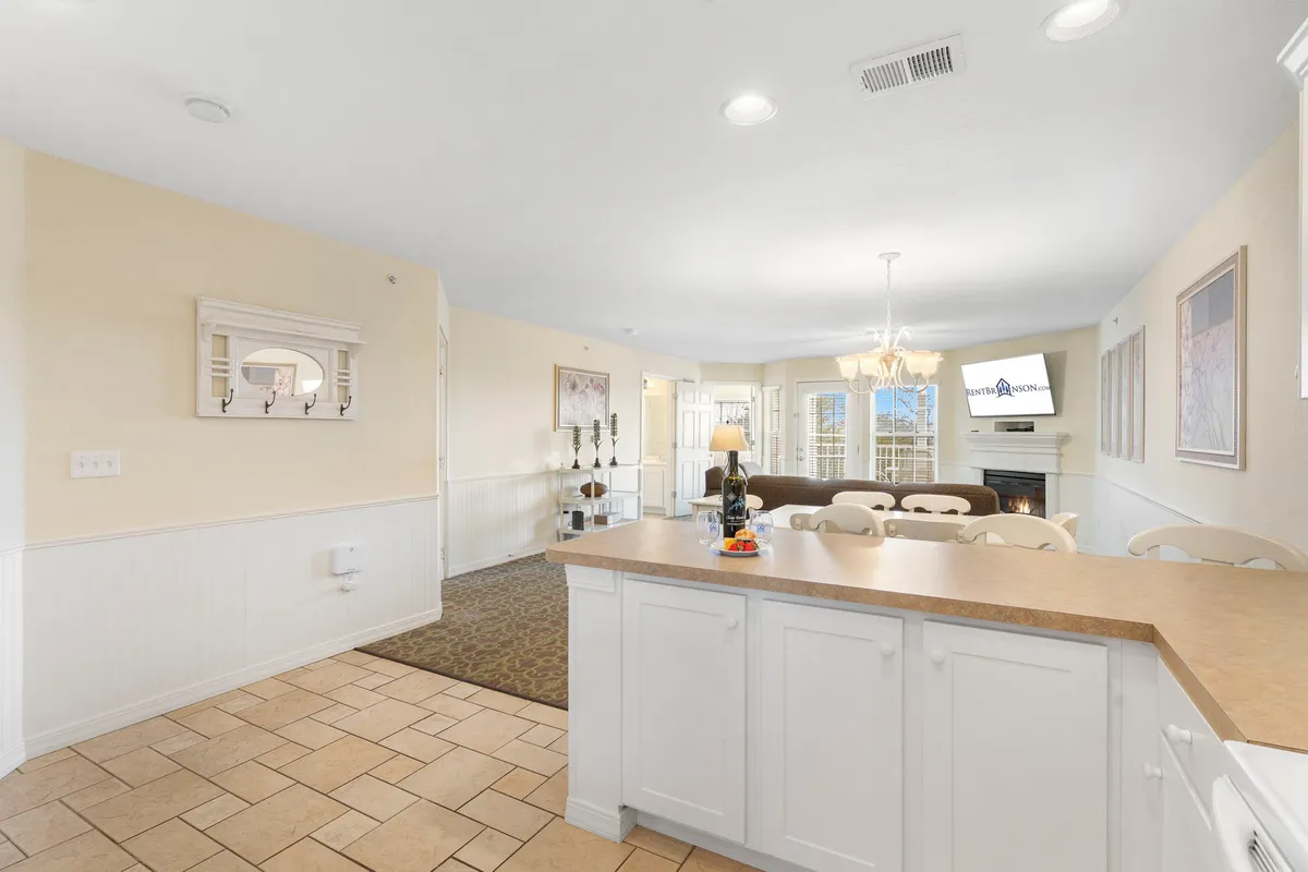 A spacious, light-filled kitchen and dining area featuring a central island, elegant chandelier, fireplace, and ample seating for family meals. Large windows flood the room with natural light, emphasizing the kitchen's inviting and functional layout.