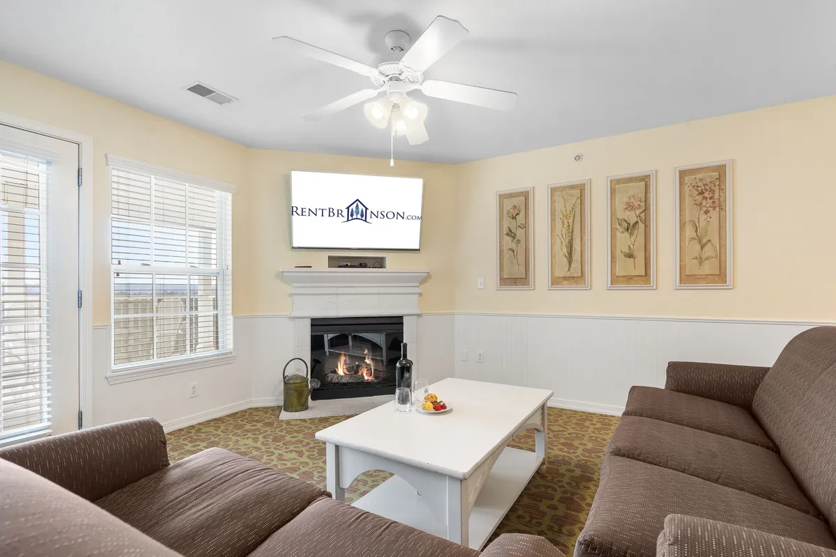 A comfortable living room anchored by a plush sofa, coffee table, and a prominent fireplace, accented by framed floral artwork. The space is designed for relaxing, watching TV, or enjoying quiet moments, all bathed in soft light from adjacent windows.