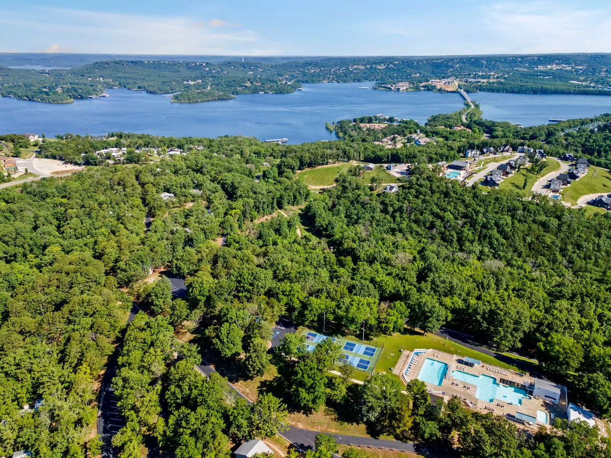 A sweeping view of Table Rock Lake with the resort's swimming pool in the foreground, surrounded by lush greenery and residential buildings. The image captures both the lakeside setting and the pool, with people enjoying swimming, hiking trails, and family gatherings visible in the scene.